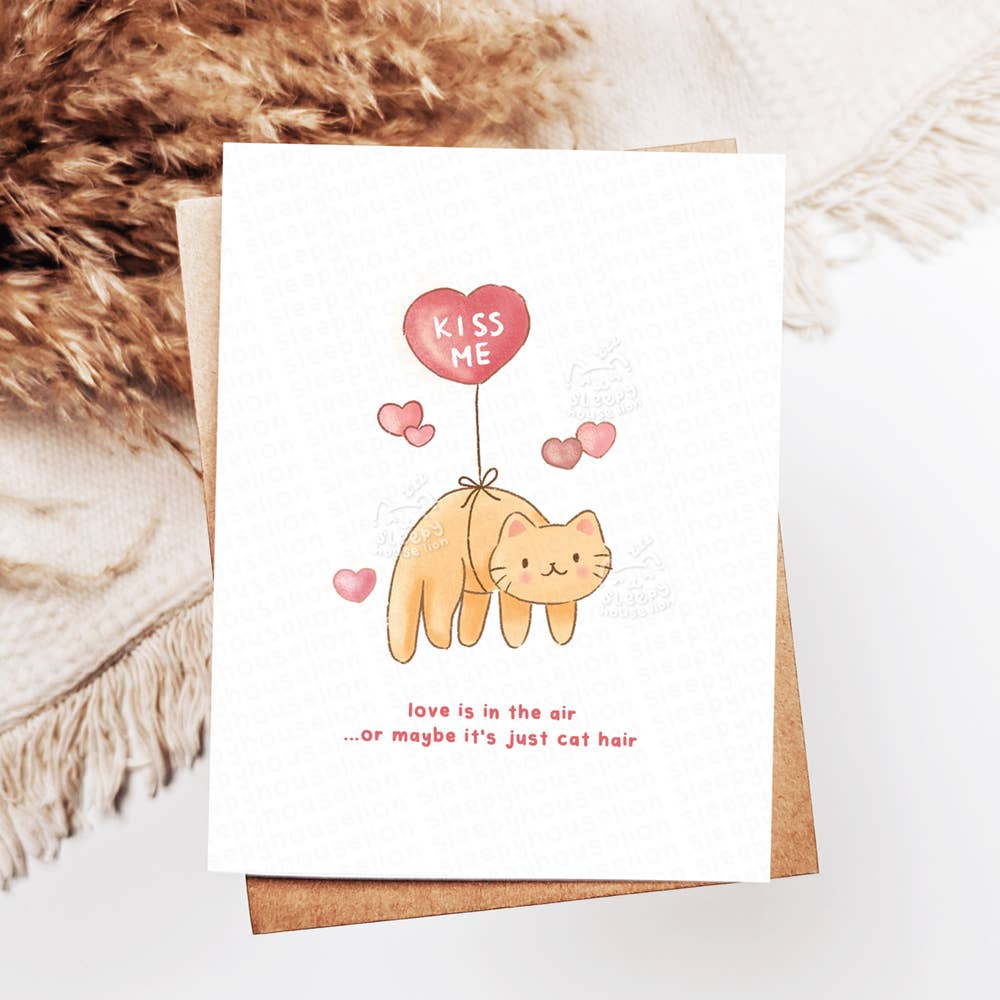 Greeting card with a cat illustration and 'Kiss Me' balloon on a textured surface.