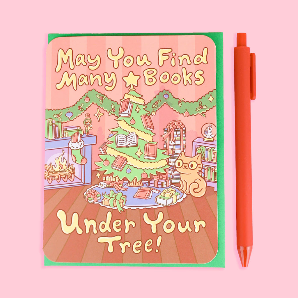 Card with Christmas illustration and text "May You Find Many Books Under Your Tree!" on a pink background with a red pen beside it