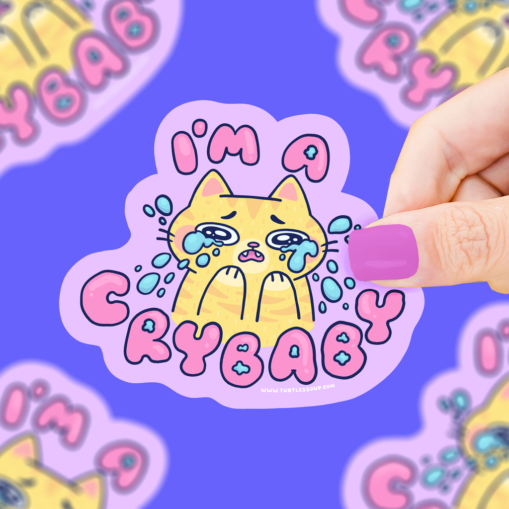 Sticker with a cat illustration and 'I'm a Crybaby' text held by a hand against a colorful background.